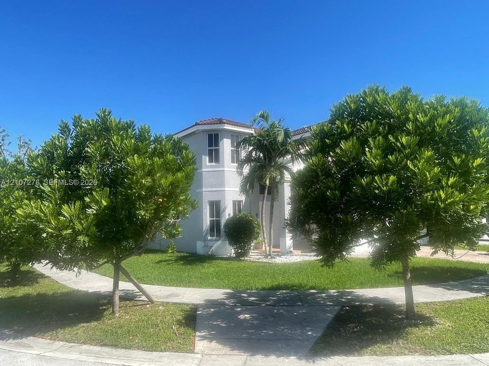 Two-story exterior featuring a light-toned stucco facade, a multi-pane window configuration, a tile roof, and a mature landscaped yard with concrete walkways