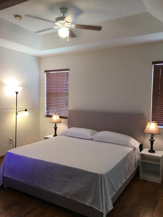 Tray ceiling, white ceiling fan with integrated lighting, two windows with horizontal blinds, and wood-finish flooring