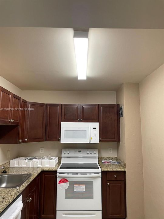 Kitchen featuring dark wood cabinetry, stone-finish countertops, stainless steel sink, and white appliances including a built-in microwave and range