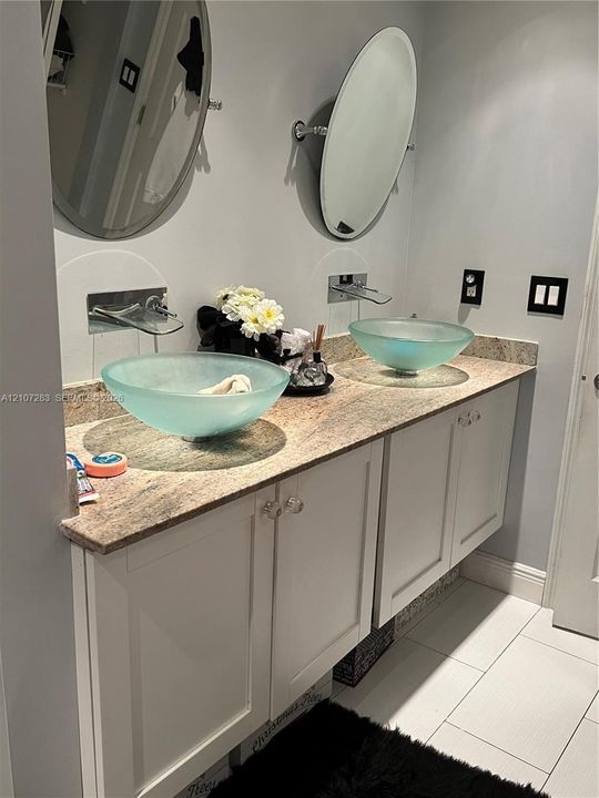 Dual vanity featuring white shaker-style cabinetry, a stone countertop, wall-mounted faucets, frosted glass vessel sinks, and oval mirrors