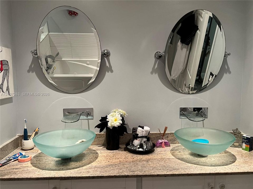 Dual vanity featuring frosted glass vessel sinks, stone countertop, wall-mounted faucets, and oval tilting mirrors