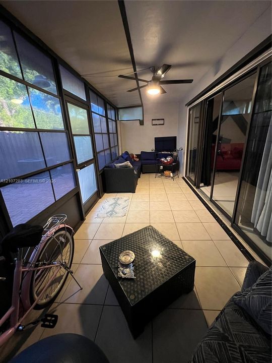 Enclosed patio featuring grid-patterned window panels, large-format tile flooring, sliding glass door entry, and a ceiling-mounted light fixture with a fan
