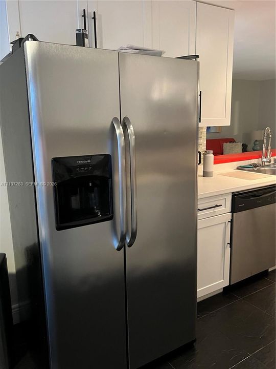Stainless steel side-by-side refrigerator, white shaker-style cabinetry with matte black hardware, stone-finish countertops, undermount sink, and dark tile flooring