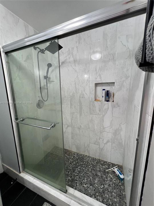 Walk-in shower featuring glass sliding doors, large-format tile surround, integrated tiled niche, rainfall shower head with handheld attachment, and a pebble-stone floor