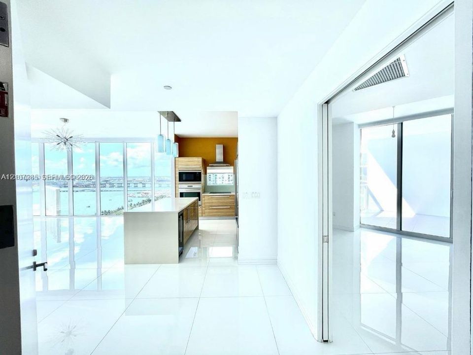 Open-concept kitchen featuring light wood-finish cabinetry, a central island with a stone countertop, built-in stainless steel appliances, white tile flooring, and floor-to-ceiling windows