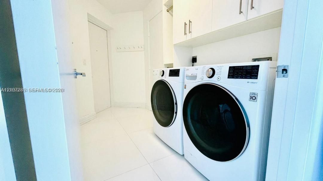 Laundry area featuring white large-format tile flooring, built-in upper cabinetry with brushed metal hardware, and a side-by-side front-loading washer and dryer set