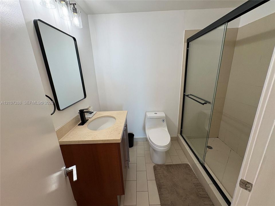 Wood-finish vanity with a light-toned countertop, undermount sink, matte black faucet, rectangular mirror with a black frame, and a walk-in shower featuring a sliding glass enclosure