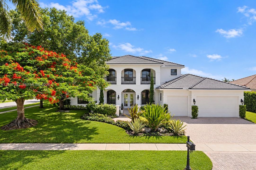 Two-story stucco exterior featuring arched windows, a covered balcony with decorative iron railings, a tile roof, and a paved stone driveway leading to an attached three-car garage