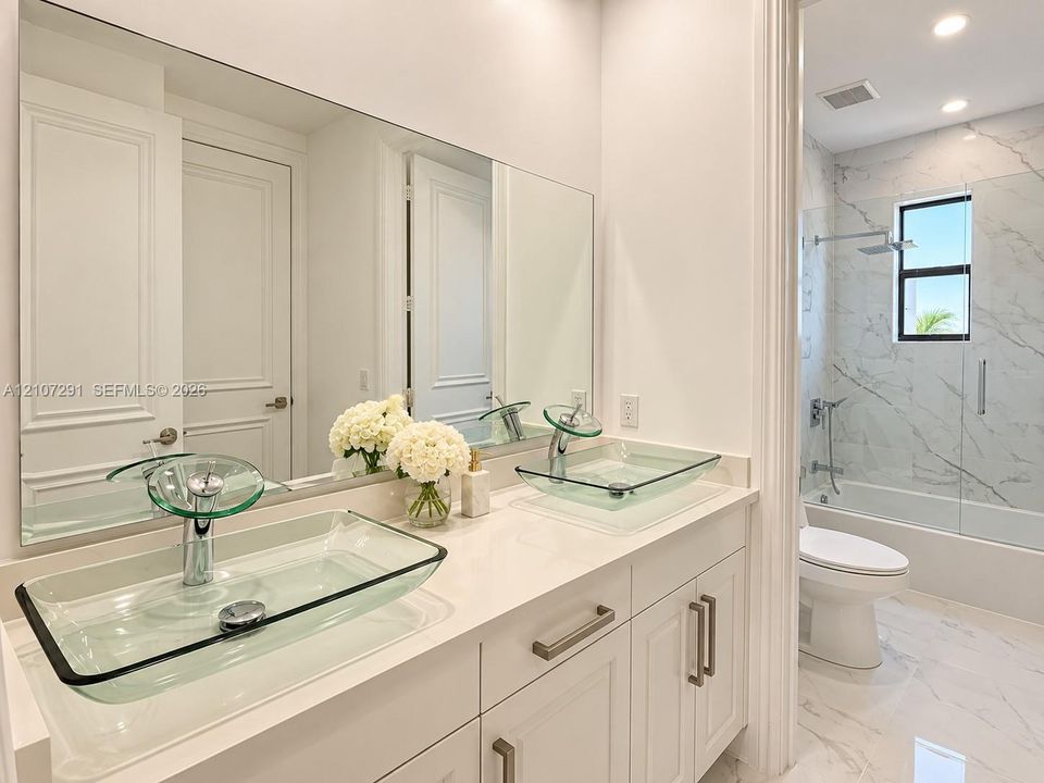 Dual glass vessel sinks featuring chrome faucets, set upon a white countertop with integrated cabinetry, complemented by a stone-tile surround in the shower and recessed lighting