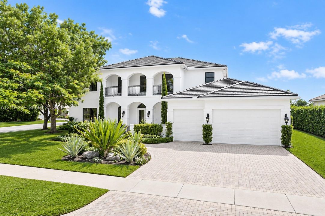 Stucco exterior featuring arched architectural detailing, a covered second-story balcony with wrought-iron railing, a paved stone driveway, a multi-car garage, and manicured landscaping