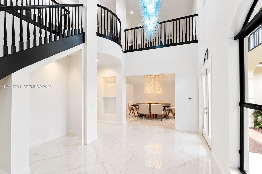 Double-height foyer with stone-finish flooring, a curved staircase featuring black iron balusters, built-in display niches, and a multi-tiered glass chandelier