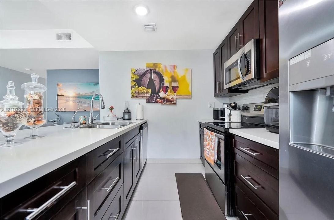 Kitchen featuring dark wood cabinetry, white countertops, stainless steel appliances, a center island with an undermount sink, and tile flooring