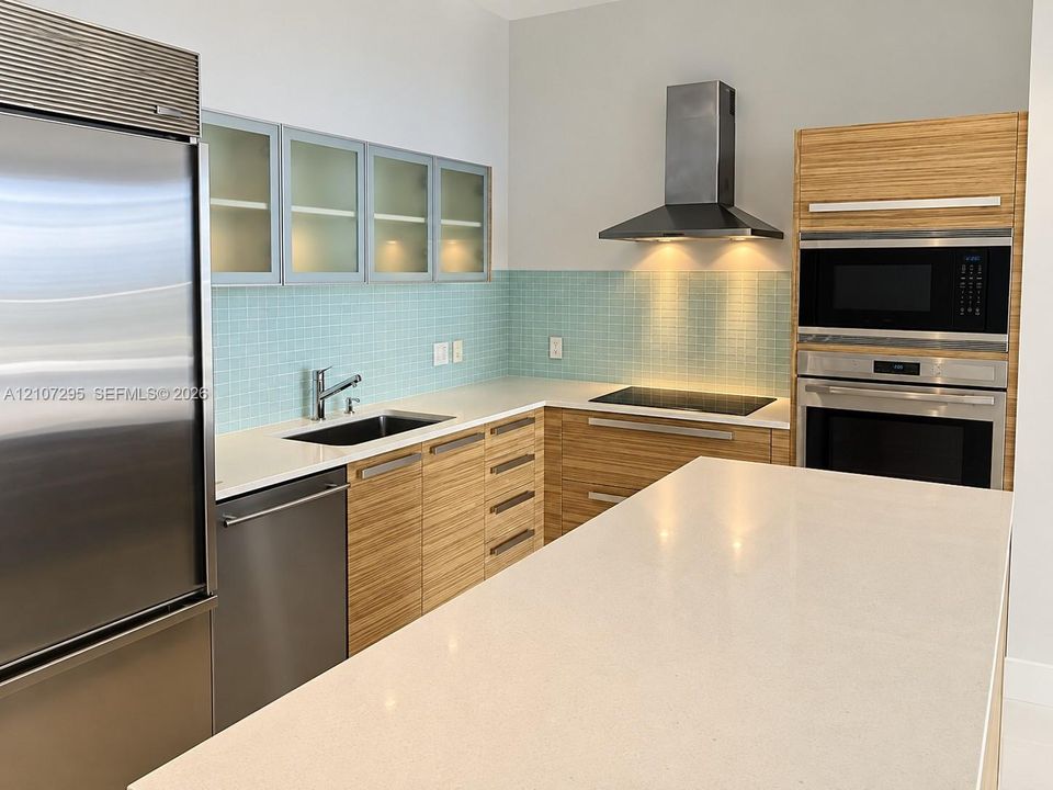 Kitchen featuring horizontal-grain wood cabinetry, stainless steel appliances, a large stone-surface island, mosaic tile backsplash, and frosted glass upper cabinet doors