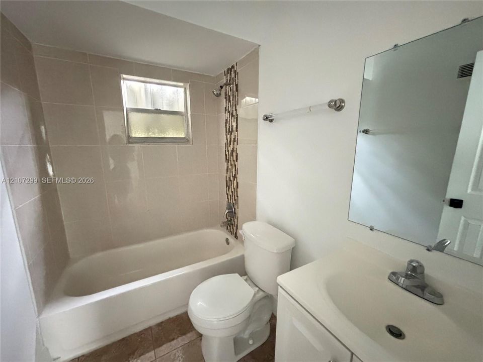 Bathroom featuring a tub with neutral tile surround, a decorative glass-tile accent strip, a white vanity with an integrated sink, and a frameless wall-mounted mirror