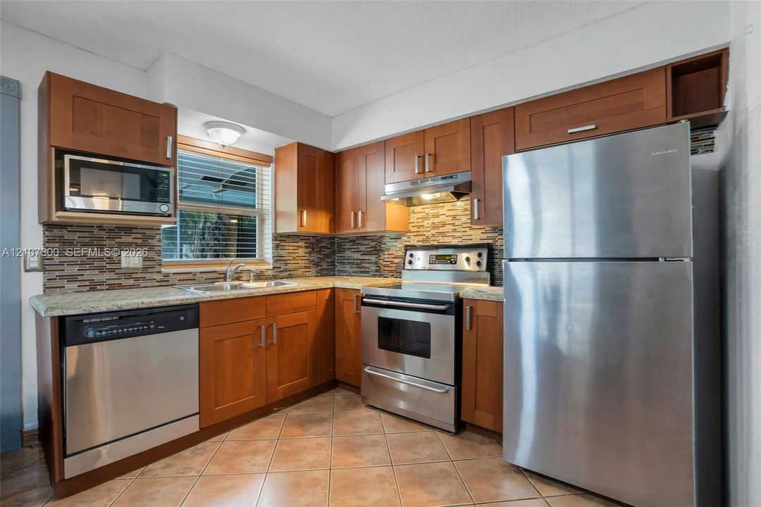Kitchen featuring wood cabinetry, stone-finish countertops, multi-toned horizontal tile backsplash, and stainless steel appliances including a range, microwave, and dishwasher