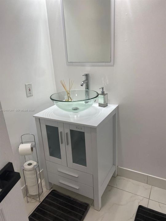 White vanity featuring cabinet storage with glass panel doors, a circular clear glass vessel sink, chrome single-handle faucet, and large-format tile flooring