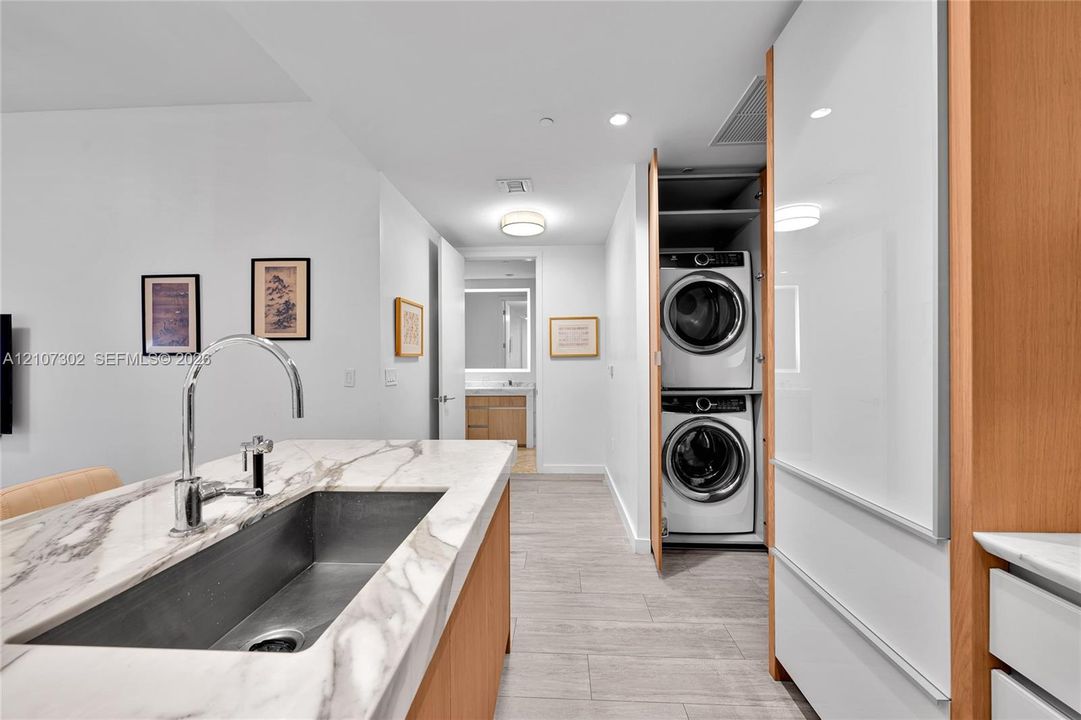 Kitchen island with stone countertop, stainless steel undermount sink, high-arc faucet, wood-finish cabinetry, and built-in laundry closet with stacked washer and dryer