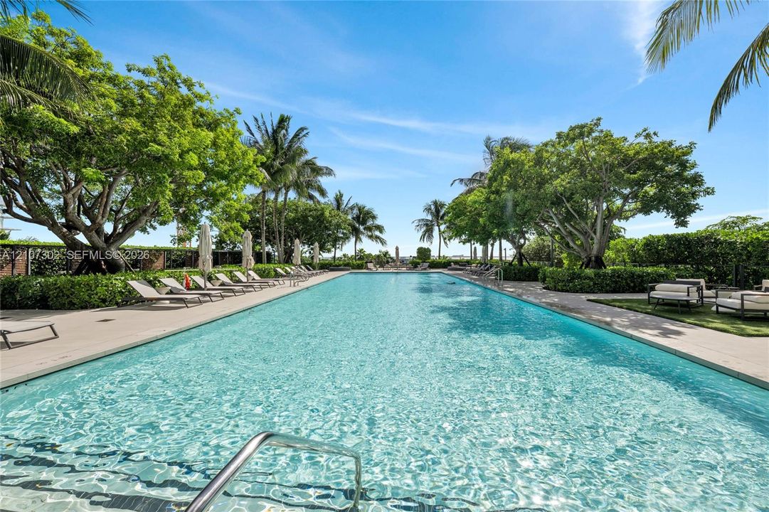 Olympic-length swimming pool featuring a concrete deck, built-in metal handrail, and surrounding mature landscaping