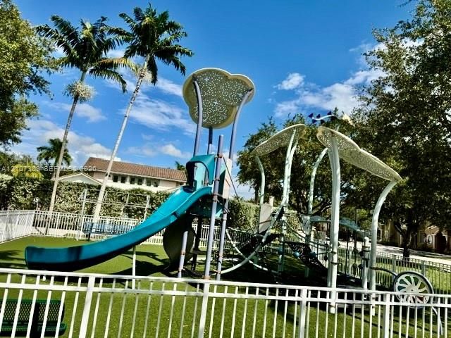 Recreational area featuring a structured playground, synthetic turf surface, metal perimeter fencing, and mature palm trees