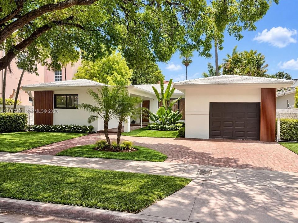 Single-story residence featuring a crisp white exterior with vertical wood-accented columns, a paved brick driveway, a dedicated garage, and mature landscaping