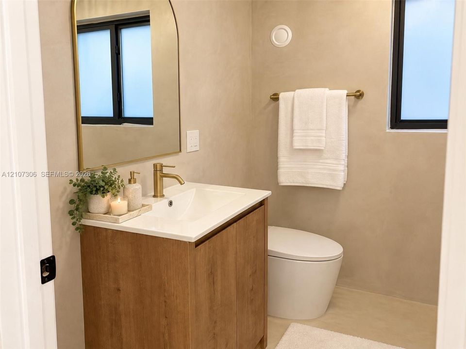 Wood-finish vanity cabinet with a white basin, arched wall mirror, brass-toned faucet, and hardware, complemented by textured wall finishes and a dual-flush toilet