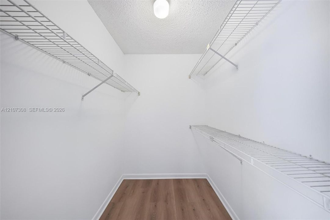 Storage area featuring wood-finish flooring, white walls, multiple tiers of white wire shelving, and a ceiling-mounted light fixture