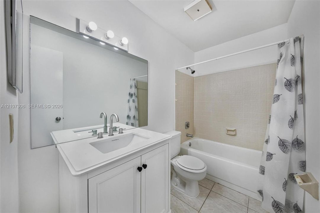White vanity cabinet with an integrated rectangular sink and polished chrome faucet, large wall-mounted mirror with three-bulb vanity light, and a tub-shower combination featuring tile surround