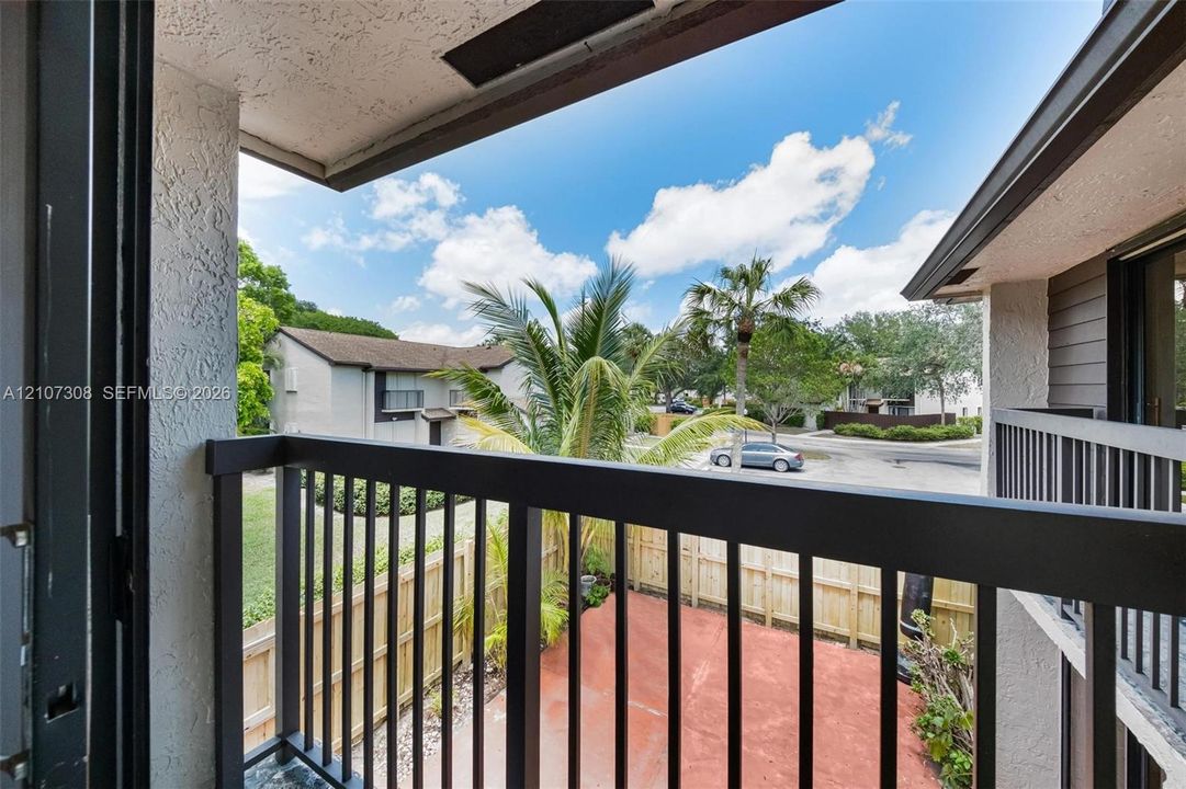 Covered balcony with a dark metal railing, overlooking a fenced ground-level patio and mature palm landscaping