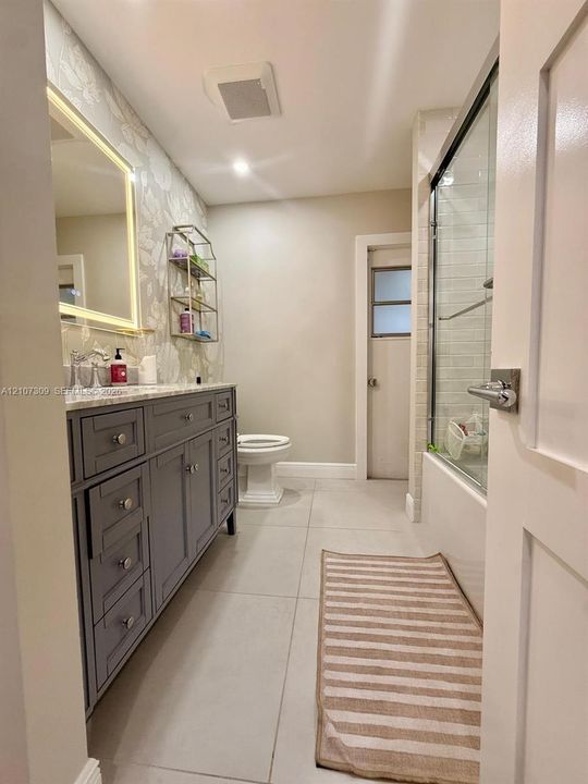 Bathroom featuring a grey shaker-style vanity with stone-surface countertop, LED-backlit mirror, large-format floor tile, and a shower-tub combination with glass enclosure