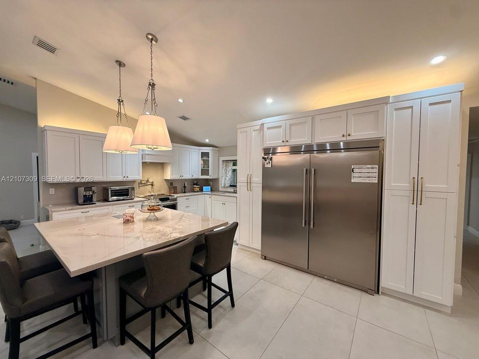 Kitchen featuring white shaker-style cabinetry, a large central island with light-toned stone countertop, stainless steel refrigerator with custom panel-matched cabinetry surround, and dual pendant lighting