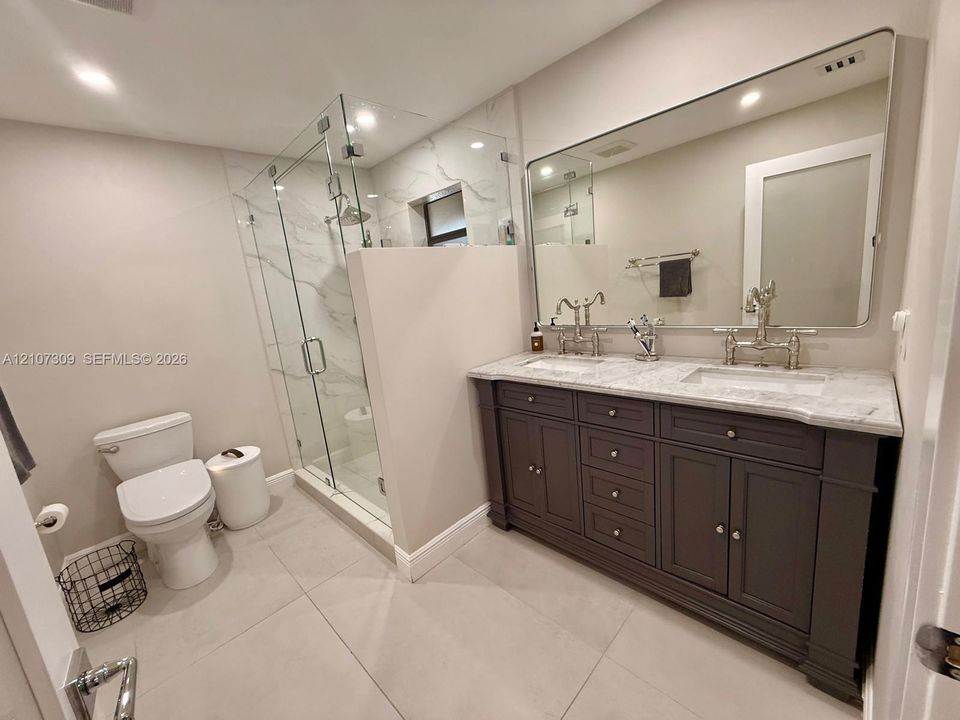 Dual-sink vanity with stone countertop and metallic fixtures, large wall-mounted mirror, walk-in glass-enclosed shower featuring stone-finish wall tile, and large-format floor tile
