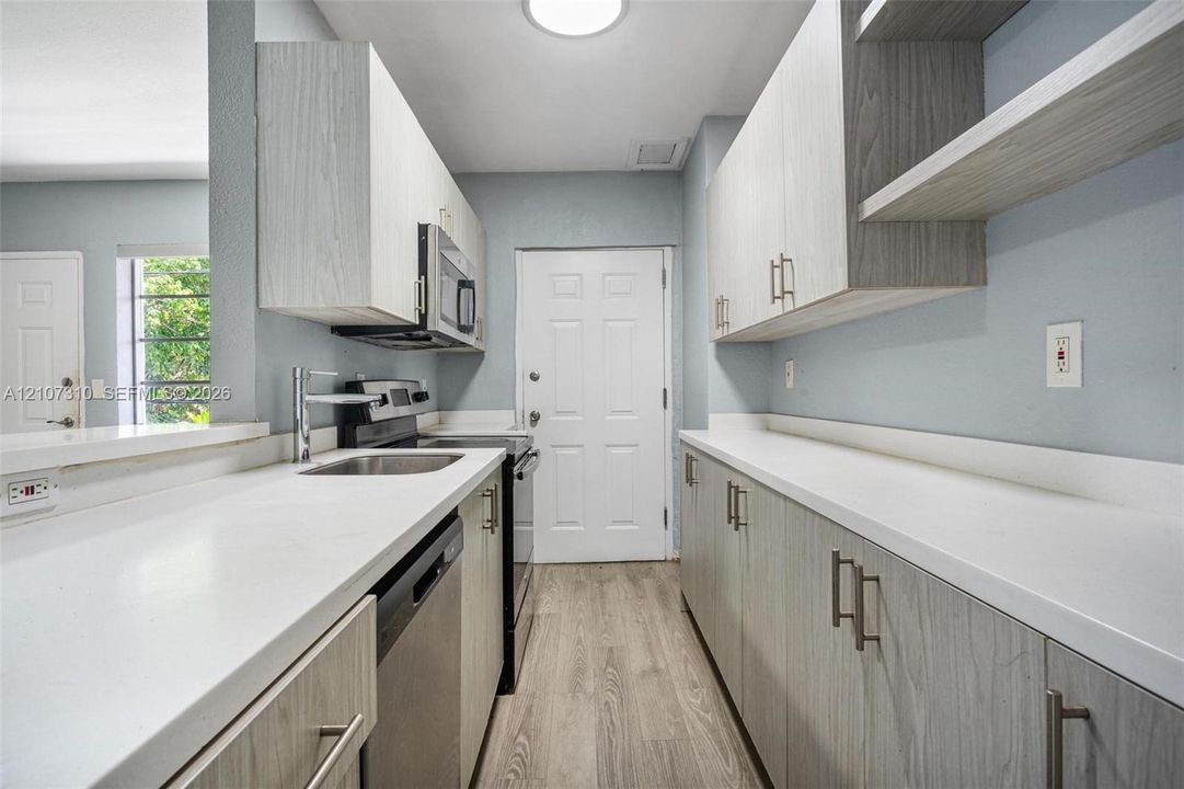 Galley kitchen featuring light-toned wood-finish cabinetry, solid surface countertops, stainless steel appliances, and wood-finish flooring