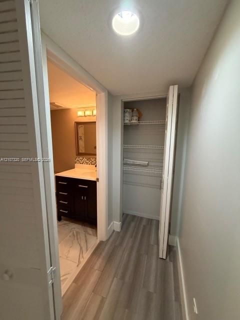 Storage closet with multi-tier wire shelving, wood-finish flooring, and recessed ceiling lighting, revealing a bathroom vanity with dark wood cabinetry and stone-finish flooring