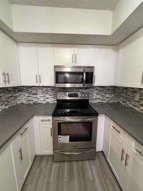 Kitchen featuring white shaker-style cabinetry, stainless steel appliances, dark stone-finish countertops, multi-toned horizontal tile backsplash, and wood-finish flooring