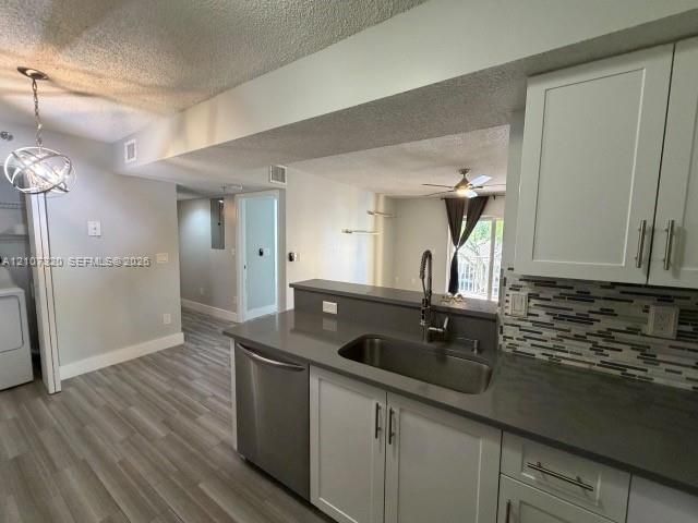 Kitchen featuring white shaker-style cabinetry, solid-surface countertops, a stainless steel undermount sink with high-arc pull-down faucet, and a multi-toned linear tile backsplash, set against wood-finish flooring