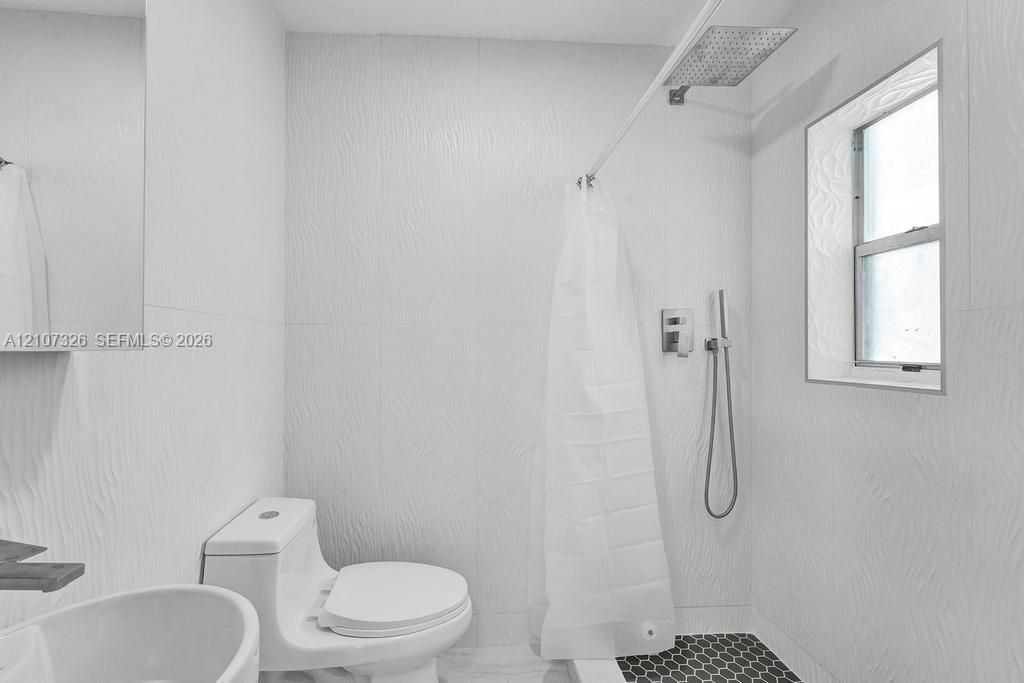 Bathroom featuring textured wall tile, rain-style shower head with handheld attachment, hexagonal floor tile, and a one-piece toilet