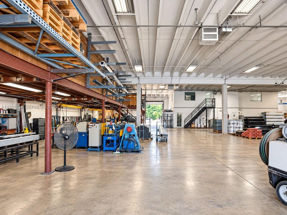 Industrial warehouse space featuring expansive concrete flooring, soaring exposed-beam ceilings, integrated structural steel mezzanine racking, and a metal staircase