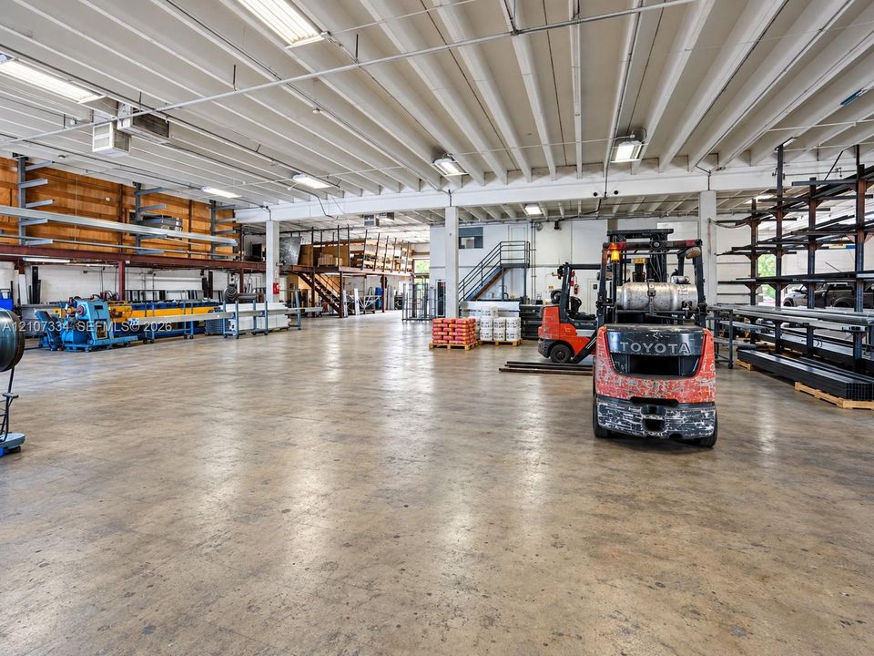 Industrial warehouse featuring high-clearance open floor space, concrete flooring, exposed overhead joists, integrated fluorescent lighting, and fixed wall-mounted steel shelving units