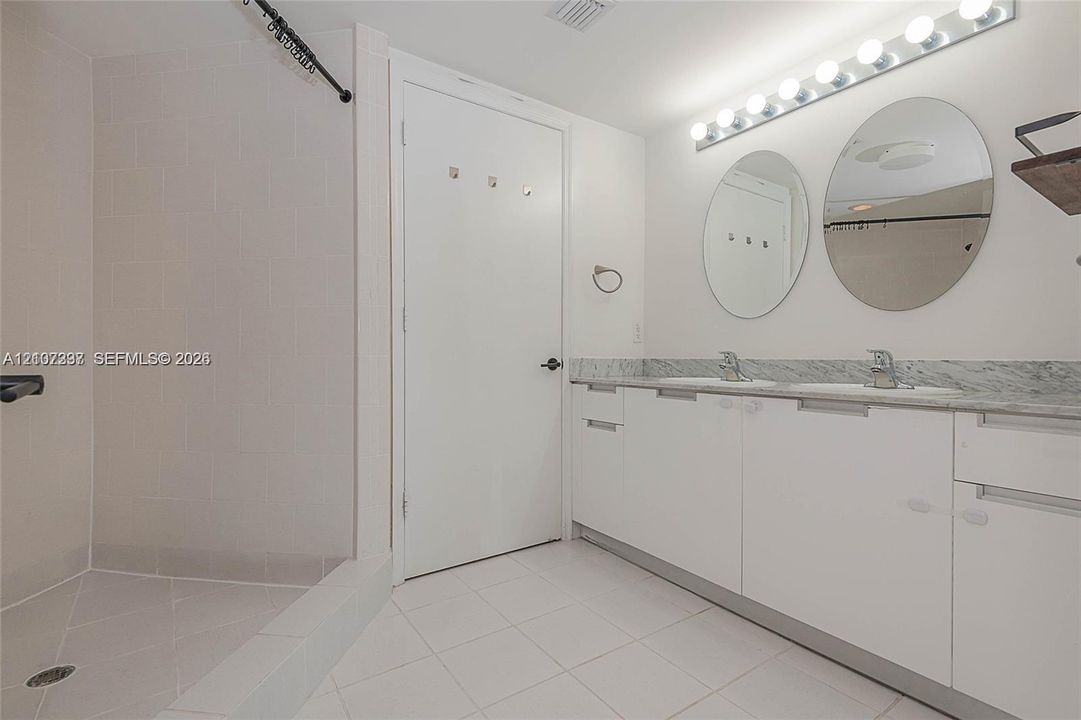 Bathroom featuring a dual-sink vanity with stone countertop, twin circular mirrors, a multi-bulb light fixture, and a walk-in shower with tile surround
