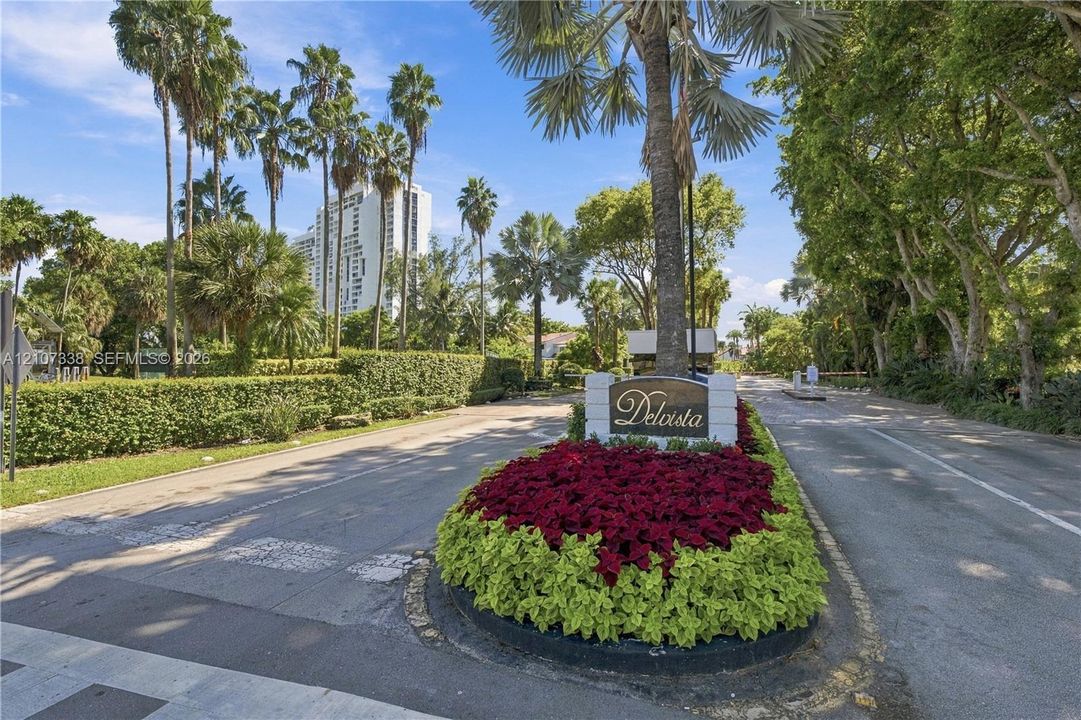 Community entrance featuring a monument sign, landscaped island with decorative plantings, palm trees, and mature canopy trees