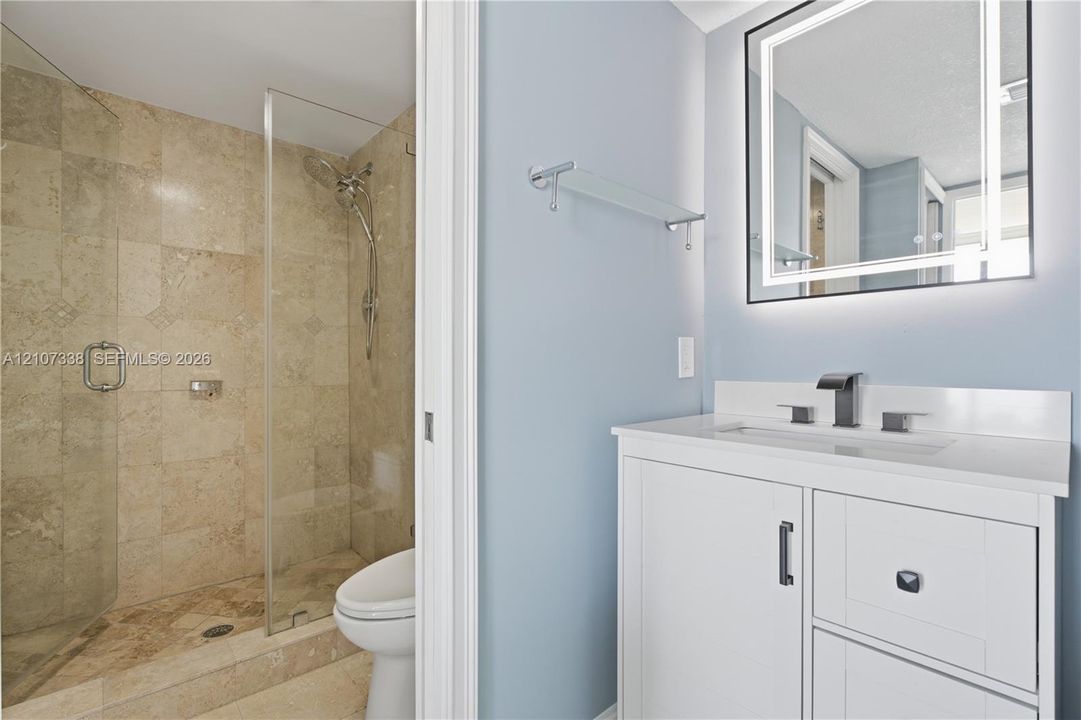 Bathroom featuring a white vanity with a quartz countertop, rectangular under-mount sink, matte black widespread faucet, and illuminated vanity mirror, alongside a walk-in shower with stone-tile surround and a glass door