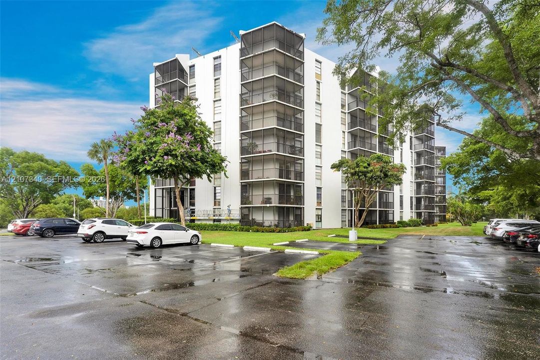 Multi-story building exterior featuring white concrete facade, vertical rows of screened balconies, paved surface parking, and mature landscaping