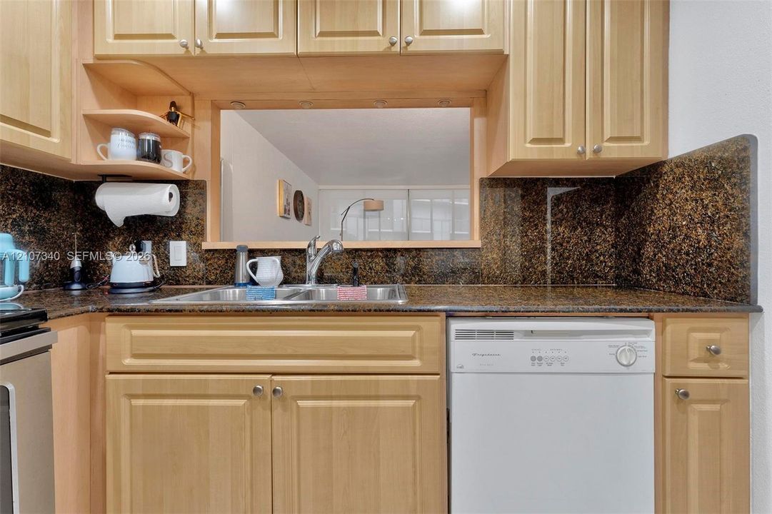 Kitchen featuring natural-finish wood cabinetry, stone-surface countertops, matching stone backsplash, dual-basin stainless steel sink with metal faucet, and built-in dishwasher