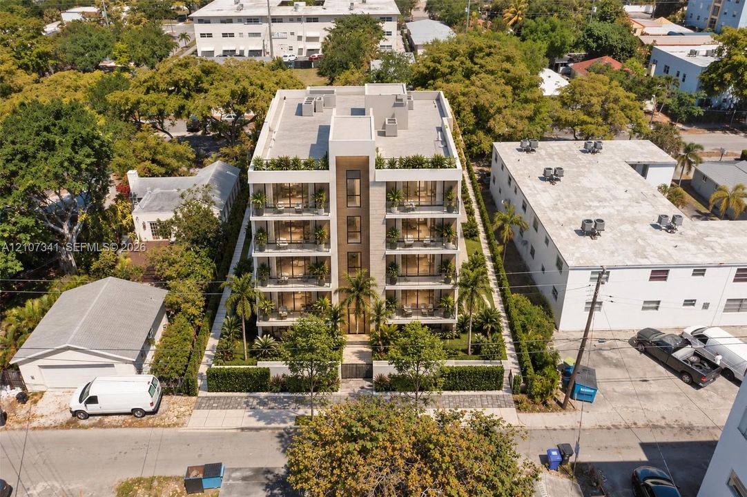 Multi-story residential building featuring a stone-clad facade, private balconies on each level, floor-to-ceiling windows, and landscaped perimeter hedging