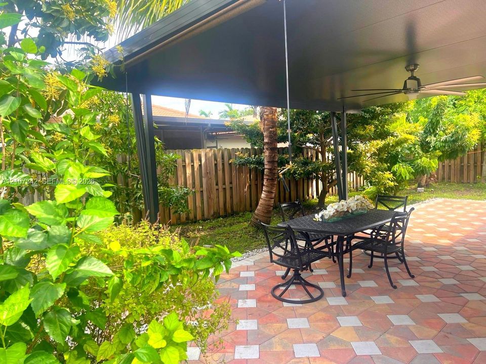 Covered patio featuring a multi-toned geometric paver floor, metal support columns, a mounted ceiling fan, and wooden perimeter fencing