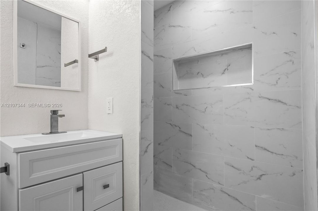 Bathroom featuring a white vanity cabinet with a rectangular undermount sink, stone-patterned tile shower surround, integrated wall niche, and metal hardware
