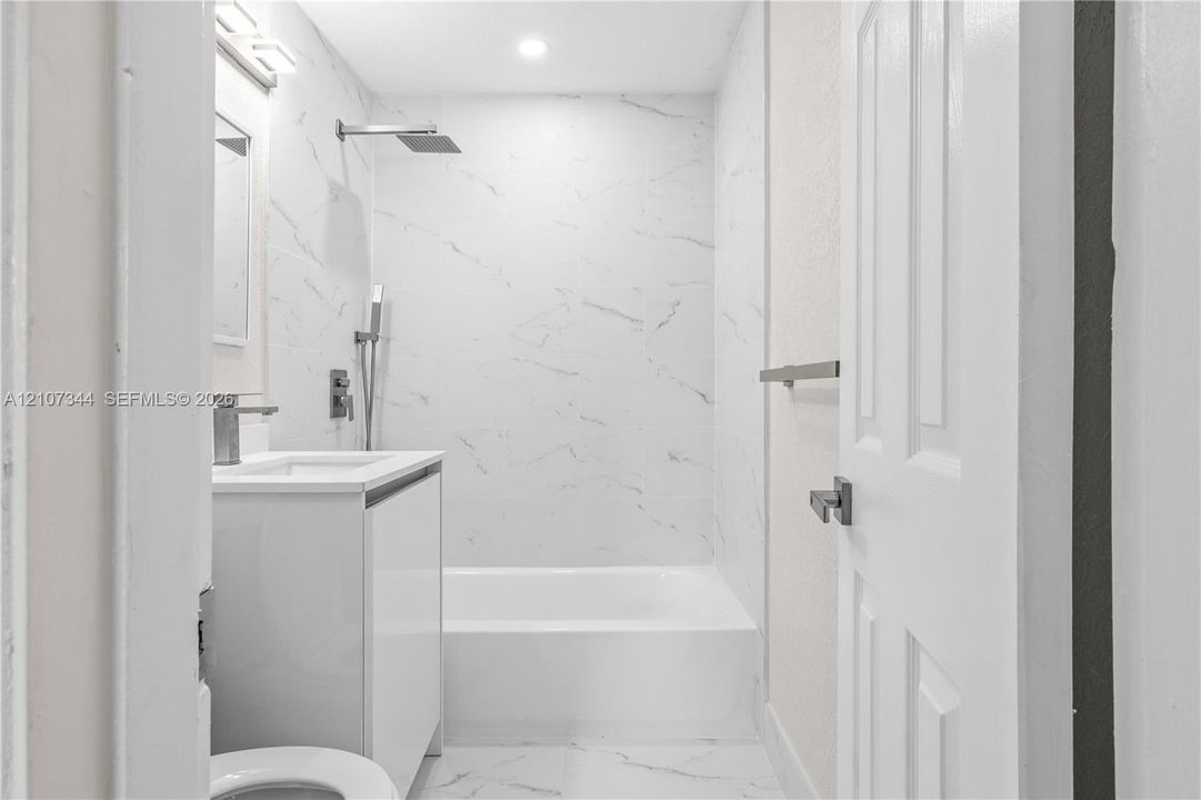 Full-height stone-finish wall tile, white vanity cabinet, rectangular undermount sink, rainfall showerhead with handheld sprayer, and recessed lighting