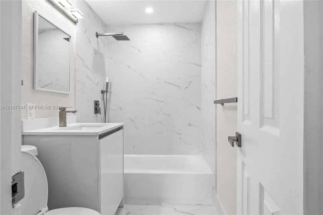 Bathroom featuring floor-to-ceiling veined stone-finish tile surround, wall-mounted rain shower head with handheld attachment, vanity with integrated basin, and matte metal hardware
