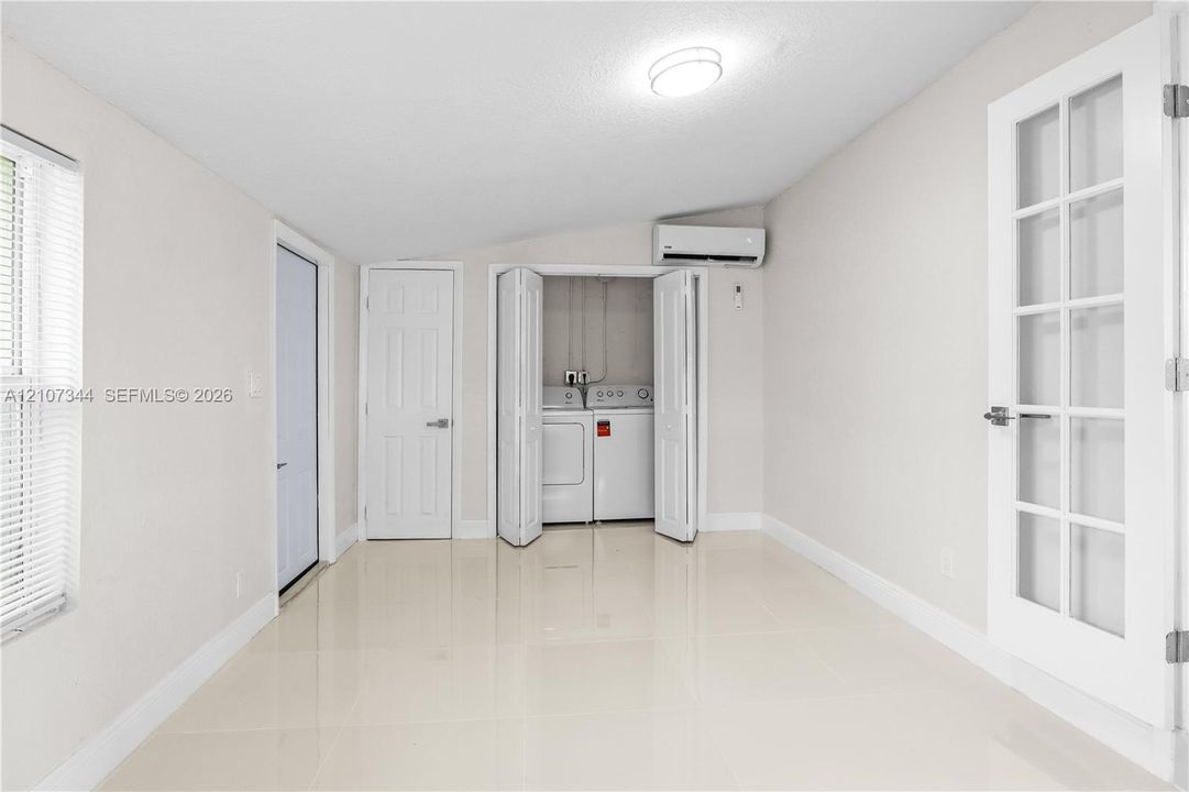 Interior space featuring light-toned tile flooring, recessed lighting, a dedicated closet with bi-fold doors containing laundry appliances, wall-mounted air conditioning unit, and a French door with glass panels