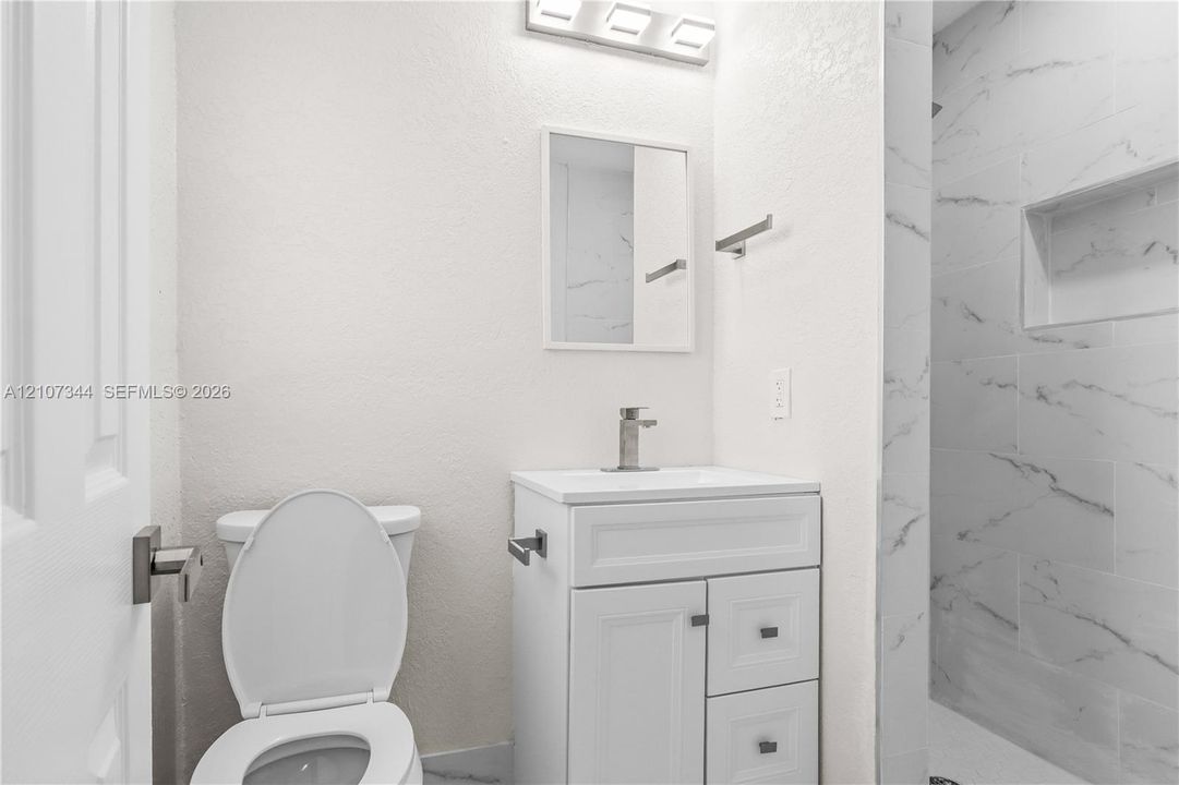 White vanity with integrated basin, polished metal faucet, wall-mounted three-light fixture, and shower enclosure featuring large-format tile with integrated recessed niche